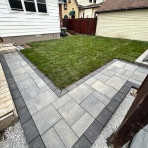 Modern backyard hardscaping featuring a sleek L-shaped paver walkway in grey tones with contrasting dark border. The clean-lined concrete pavers are laid in a structured pattern, framing a well-maintained lawn area. The walkway connects to a wooden deck and is bordered by gravel edging. White siding and wooden privacy fence complete this urban yard transformation, demonstrating professional installation and attention to detail in geometric design