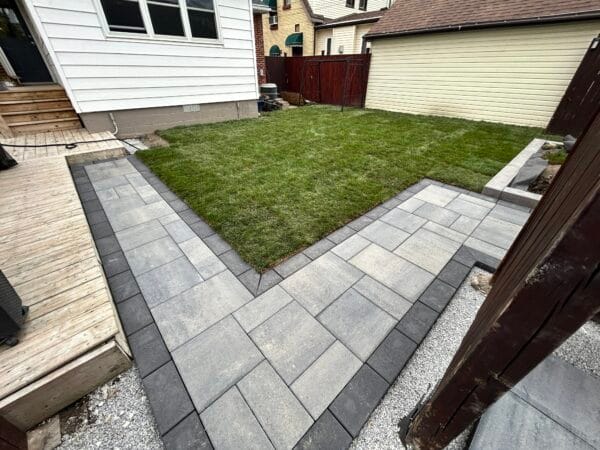 Modern backyard hardscaping featuring a sleek L-shaped paver walkway in grey tones with contrasting dark border. The clean-lined concrete pavers are laid in a structured pattern, framing a well-maintained lawn area. The walkway connects to a wooden deck and is bordered by gravel edging. White siding and wooden privacy fence complete this urban yard transformation, demonstrating professional installation and attention to detail in geometric design